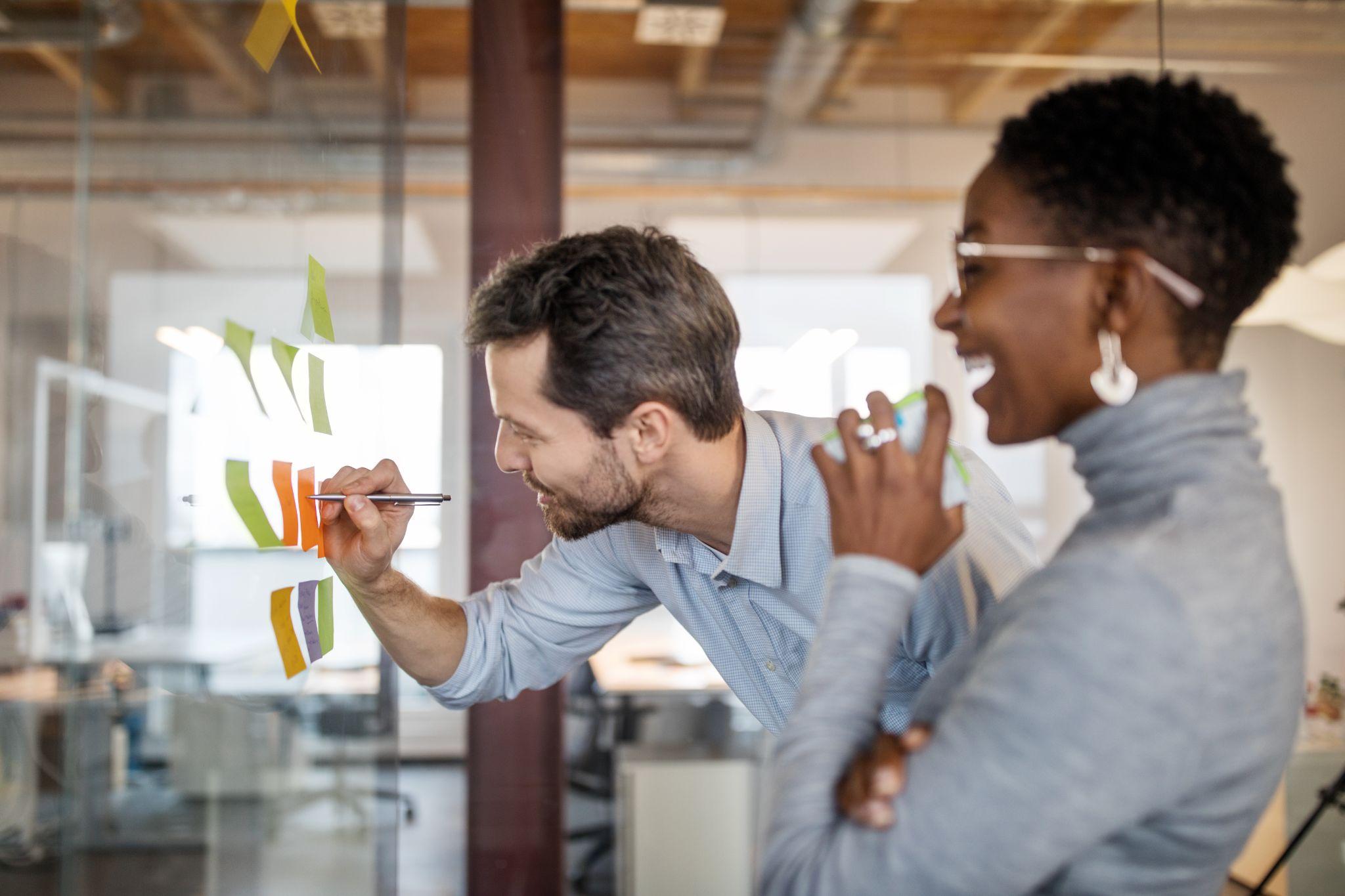 Zwei Kolleginnen und Kollegen im Workshop an einer Glaswand mit bunten Post-its bei der Strategieentwicklung