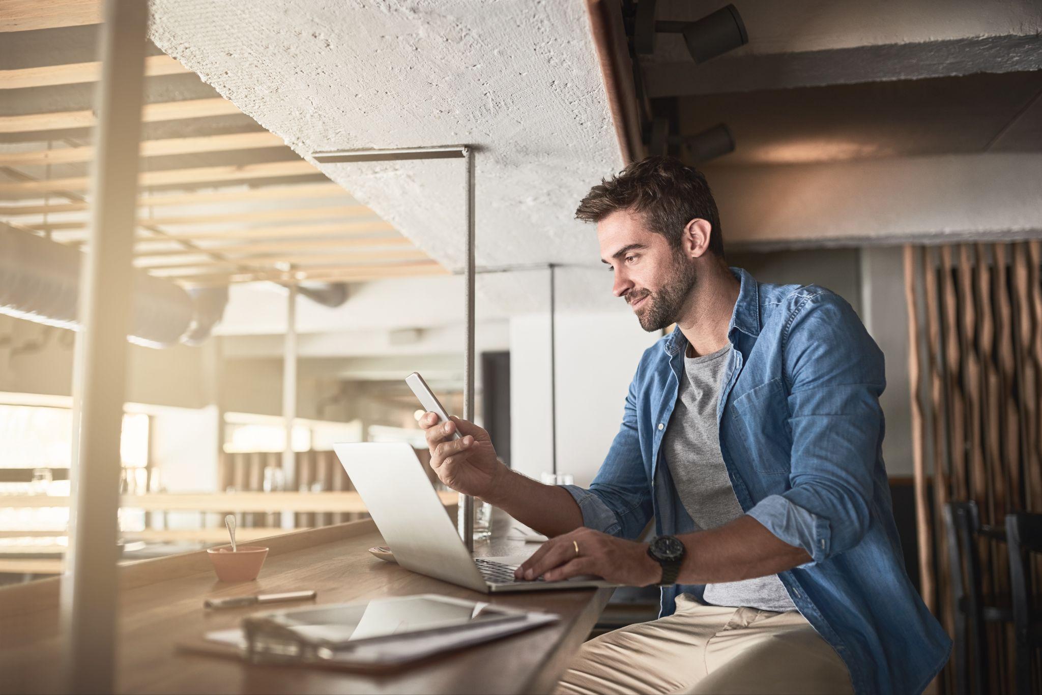 Mann arbeitet mit Smartphone und Laptop flexibel in einem Café als Beispiel für digitales Arbeiten im AXA Exklusiv-Vertrieb